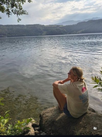 Frau im Miami-Nice-Shirt sitzt auf einem Stein am See